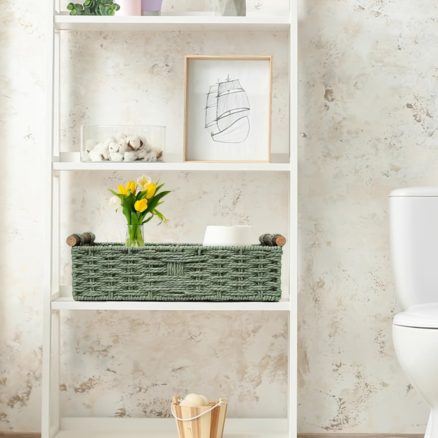 Green woven storage basket with wooden handles on a white shelf holding flowers and decorative items