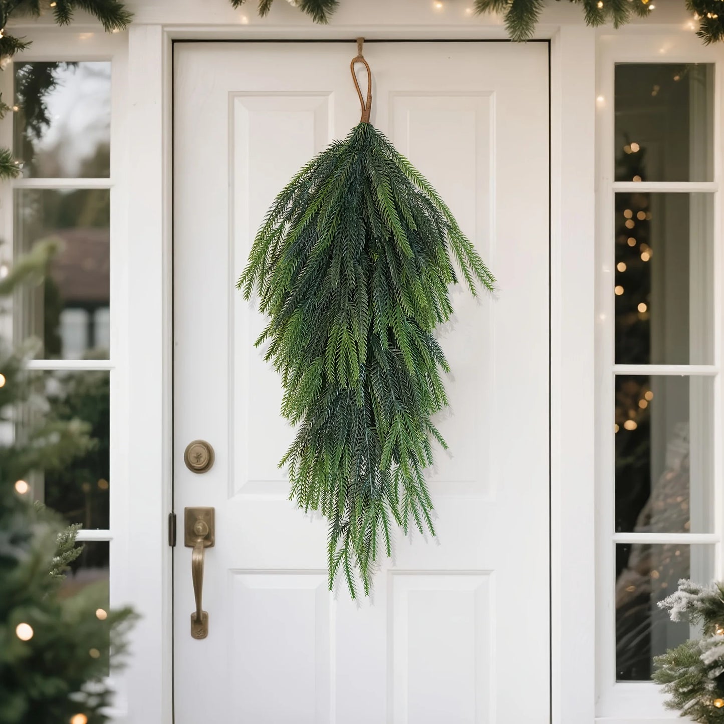 Artificial pine teardrop wreath hanging on a white front door surrounded by greenery