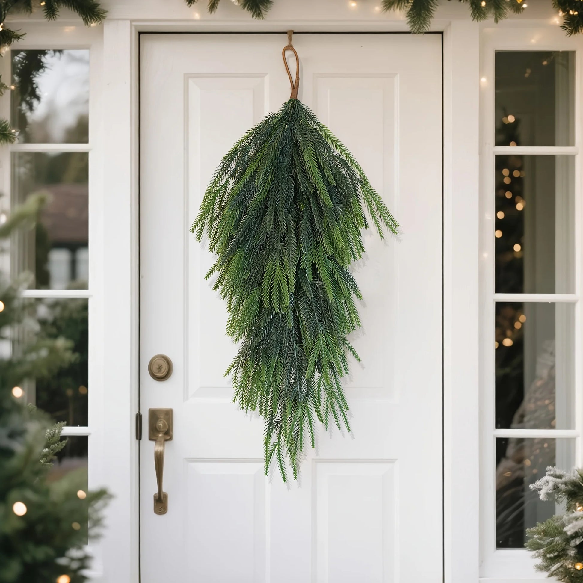 Artificial pine teardrop wreath hanging on a white front door surrounded by greenery