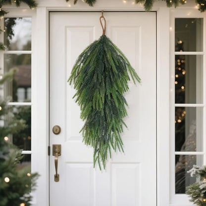 Artificial pine teardrop wreath hanging on a white front door surrounded by greenery
