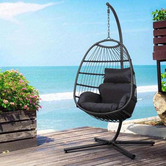 Black egg swing chair with stand on wooden deck overlooking ocean with greenery nearby