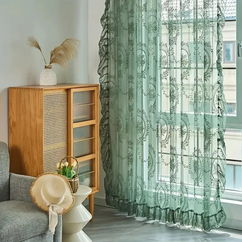 Nutshell Ruffled Floral Sheer Curtain in sage green hung in a cozy living room by window and wooden cabinet