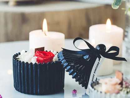Nutshell candle with wooden candle wick in black ribbed container decorated with red flower and two white candles in the background