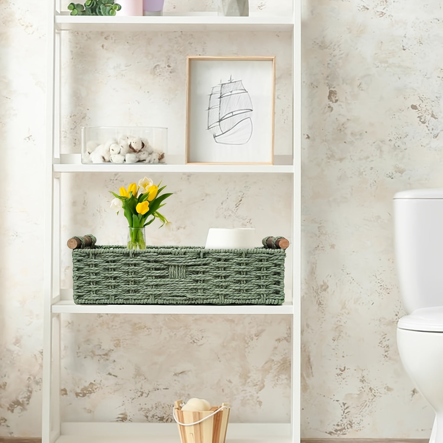 Green woven storage basket with wooden handles on a white shelf holding flowers and decorative items